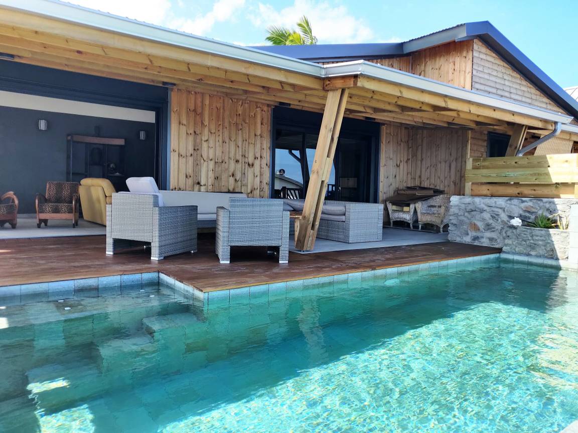 Piscine turquoise bordée du deck en bois devant la villa en bardage bois